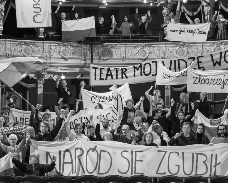 Czarno-białe zdjęcie. Grupę protestujących zgromadzonych w zdobionym wnętrzu teatru. Ludzie stoją na parterze i balkonach, trzymając transparenty oraz flagi z hasłami, wielu z nich unosi pięści w geście sprzeciwu. Na pierwszym planie widoczny jest duży baner z napisem „Naród się zgubił”.